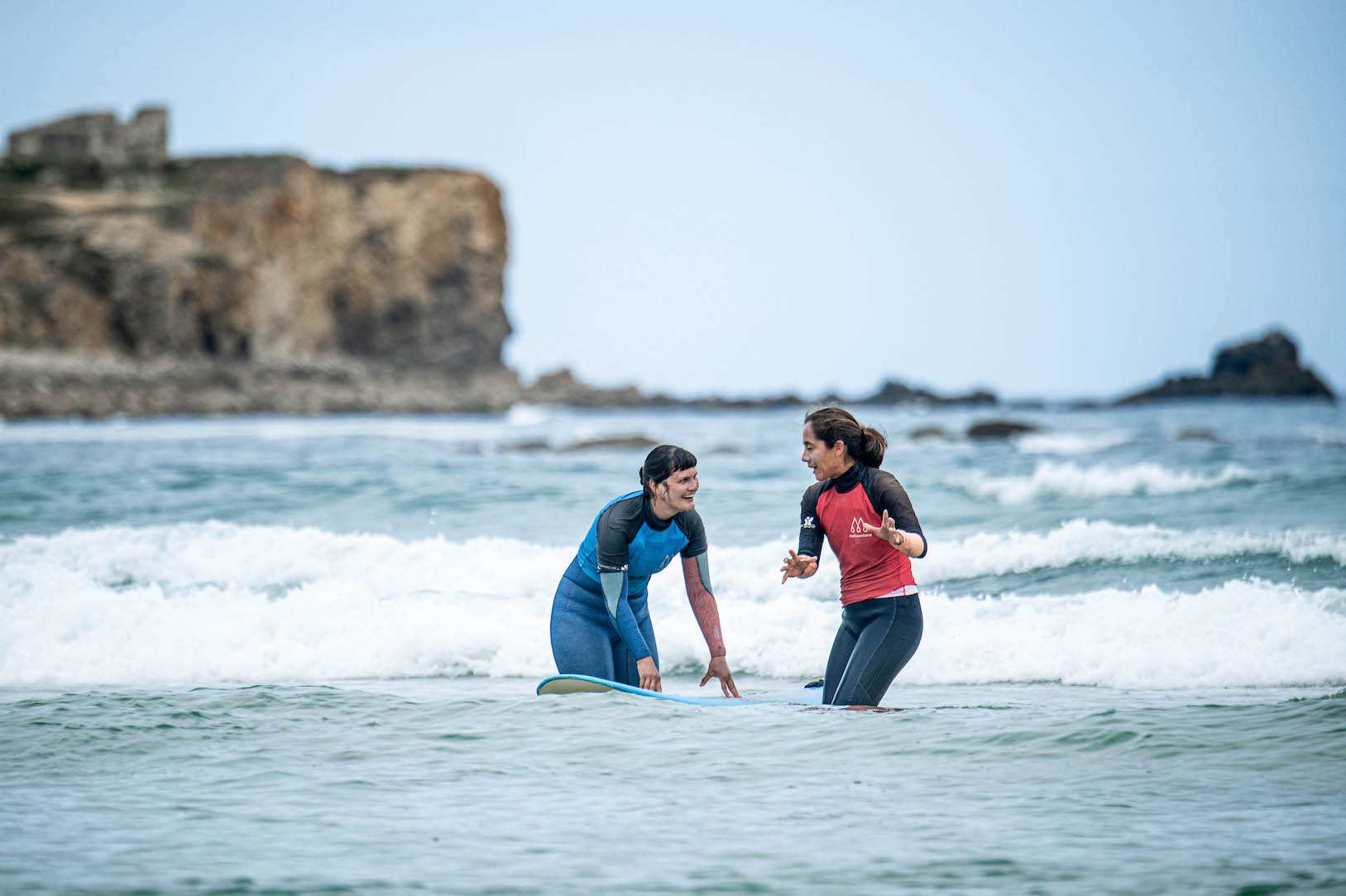 Surfcoach explaining surf technique to a student, both are standing in the the water in the ocean.
