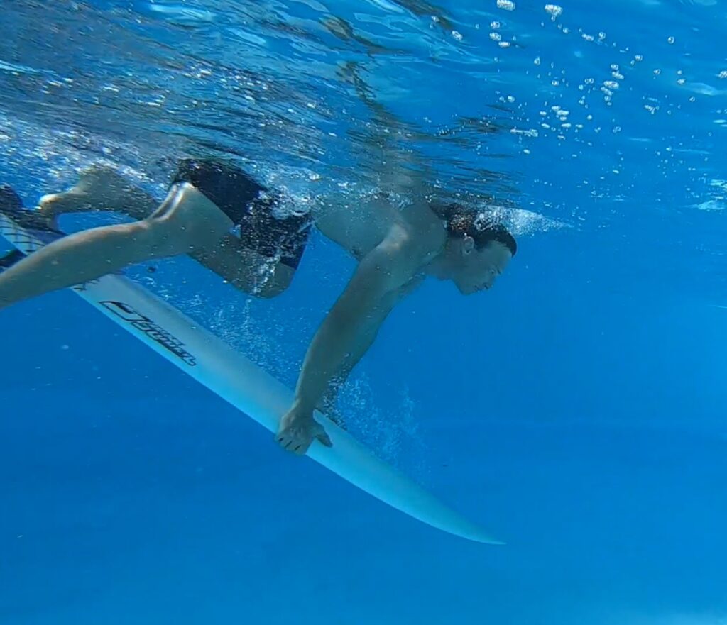 Duck Dive lernen im Pool - ein Mann auf einem Surfbrett drückt die Nase des Surfbrettes unter Wasser