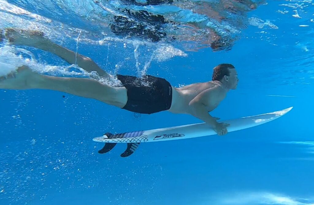 Duck Dive lernen im Pool - ein Mann drückt das Surfbrett unter Wasser und ist nun in der Auftauchphase.